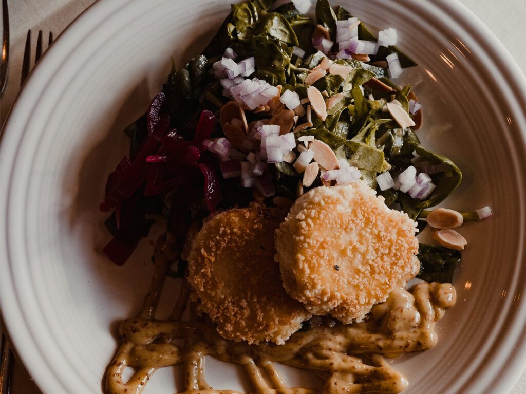 A white plate with two breaded, round patties, sautéed greens topped with chopped red onions and sliced almonds, a portion of purple pickled vegetables, and a drizzle of mustard sauce.