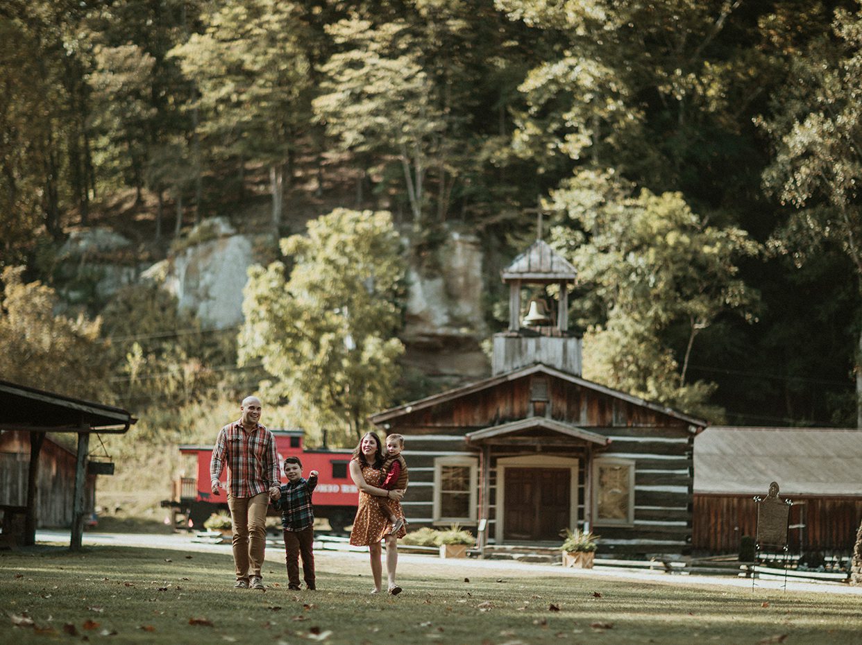 A family of four walks hand-in-hand on a grassy lawn in front of a rustic wooden building with a bell tower, surrounded by trees and greenery on a sunny day.