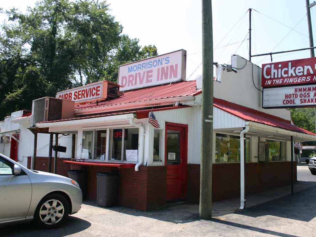 A small, retro-style drive-in restaurant with red trim, a Morrisons Drive Inn sign, and a curb service window. A car is parked in front, and menu signs are visible on the building. Trees are in the background.