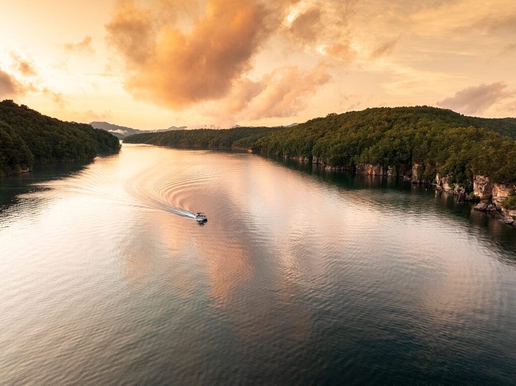 A boat travels across a calm, wide lake surrounded by forested hills at sunset, with soft clouds and the golden sky reflected in the water.