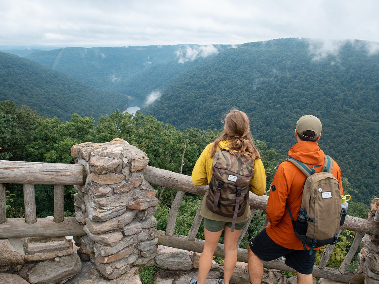 Two hikers with backpacks stand at a stone and wooden overlook, gazing at a lush, green valley with a winding river below and misty mountains in the distance.