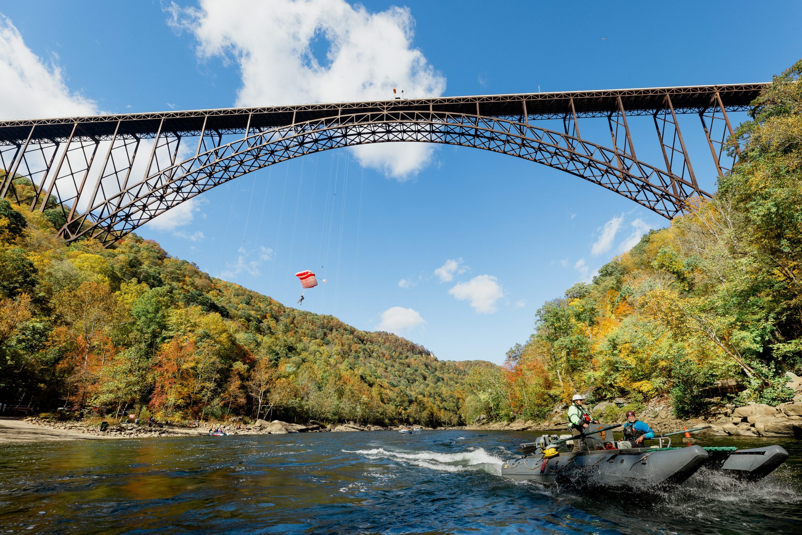 A person parachutes from a large steel arch bridge above a river, while a boat with several people speeds below; colorful autumn trees cover the surrounding hills under a blue sky.