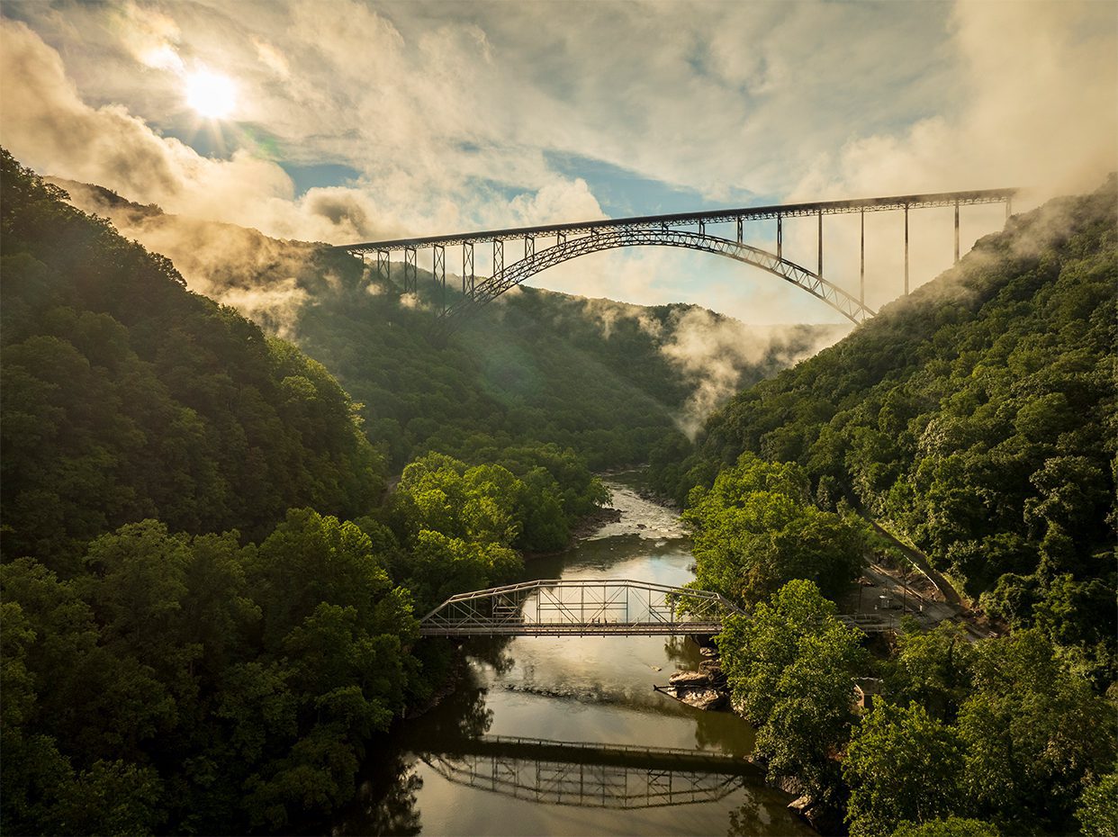 A large steel arch bridge spans a lush, green river gorge with another smaller bridge below. Mist rises from the trees, and the sun is visible through clouds in the sky.