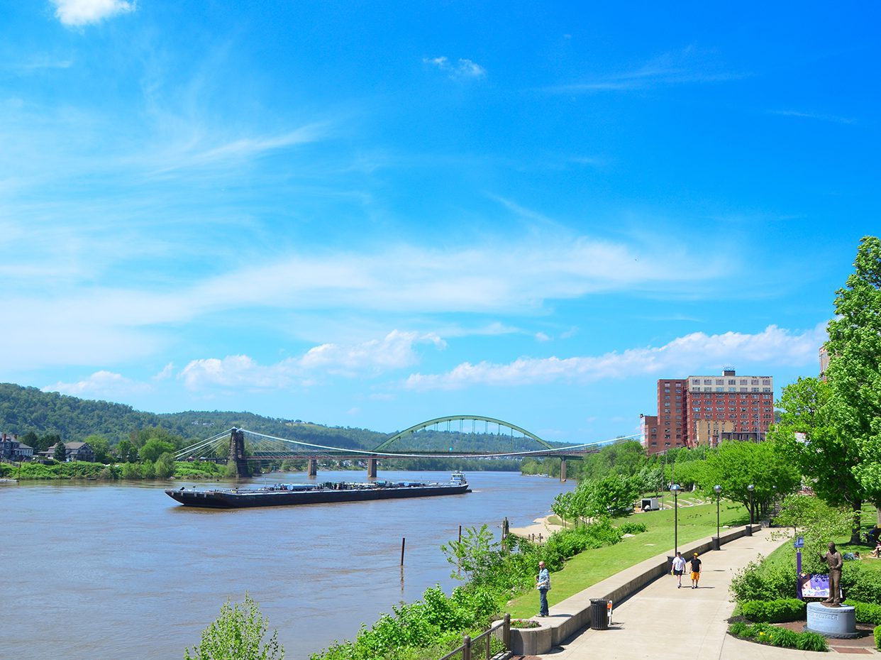 A wide river flows under a green arch bridge on a bright, sunny day. A barge travels on the water, while people walk along a paved riverside path lined with trees and a red brick building.
