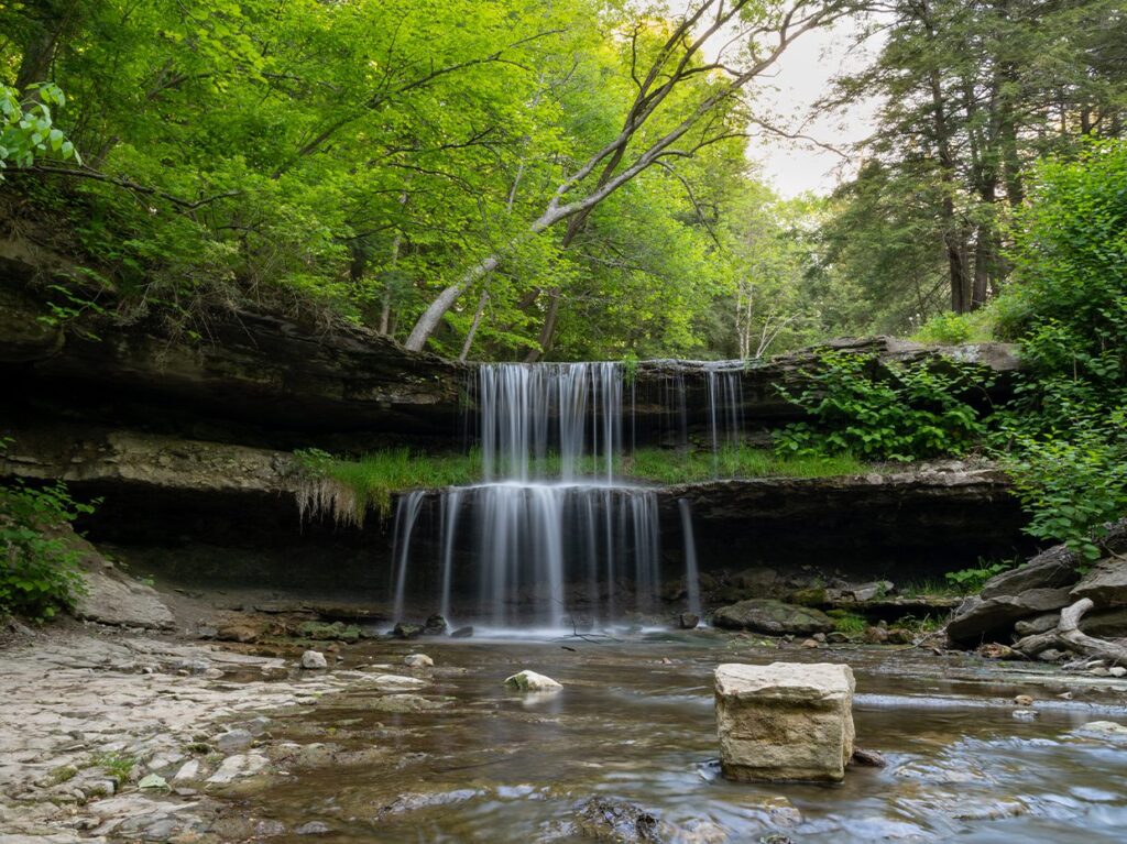 A small waterfall cascades over a rocky ledge into a shallow stream surrounded by lush green trees and foliage, with sunlight filtering through the leaves. A large stone sits in the water in the foreground.