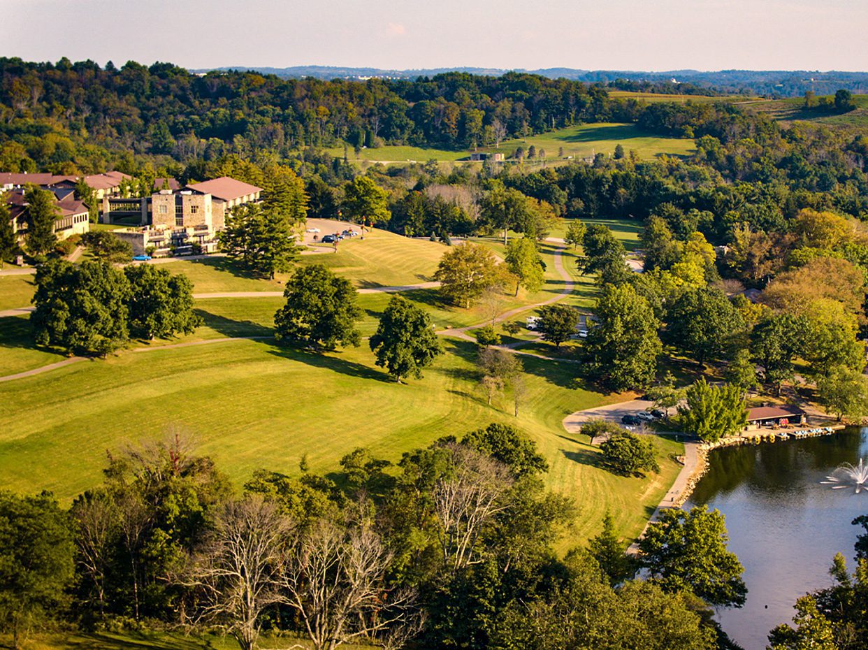 Aerial view of a lush, green landscape with rolling hills, scattered trees, a large building, winding paths, and a lake with a fountain on the right side under a clear sky.