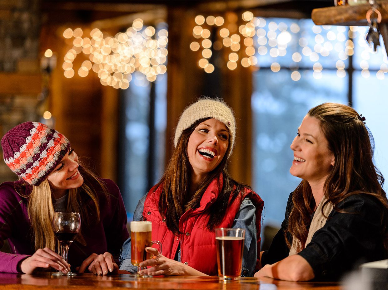 Three women wearing winter clothes sit at a wooden bar, smiling and laughing together. Drinks are on the table, and warm string lights create a cozy, festive atmosphere in the background.