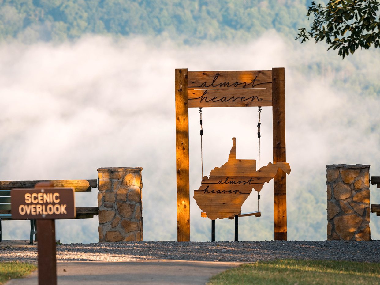 A wooden swing with a cutout of West Virginia and the words almost heaven overlooks a misty, forested valley. A sign labeled Scenic Overlook stands nearby.