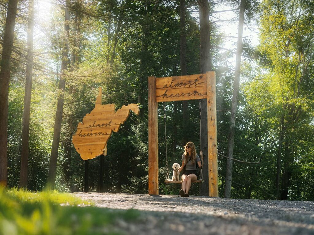 A woman sits on a wooden swing holding a small dog, surrounded by tall trees. The swing frame has the words almost heaven written on it, with a wooden cutout of West Virginia nearby. Sunlight filters through the forest.