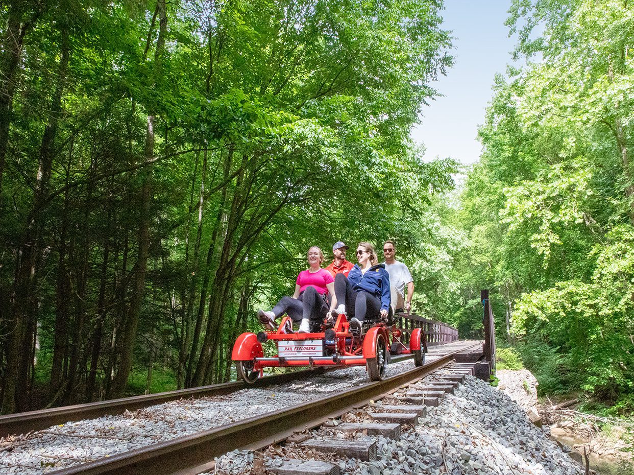 Four people ride a red rail bike along railroad tracks surrounded by lush green trees on a sunny day. The group appears to be enjoying the scenic outdoor adventure.