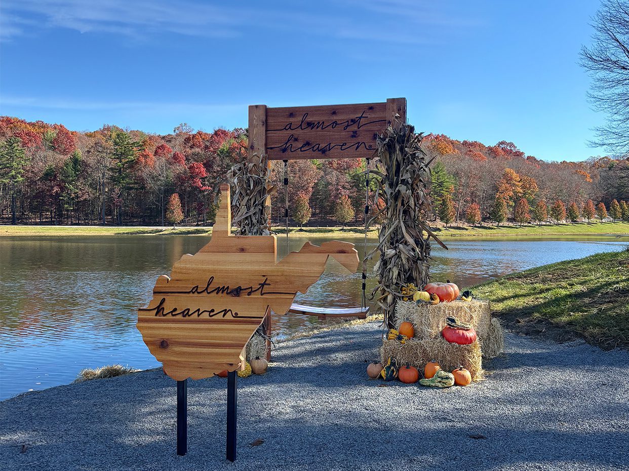 A wooden West Virginia state cutout and sign reading “almost heaven,” surrounded by autumn decorations, hay bales, pumpkins, and corn stalks by a lake with colorful fall trees in the background.