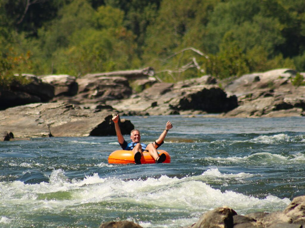 A person smiles and raises their arms while floating on an orange inner tube down a river with white water, surrounded by rocks and lush green trees.
