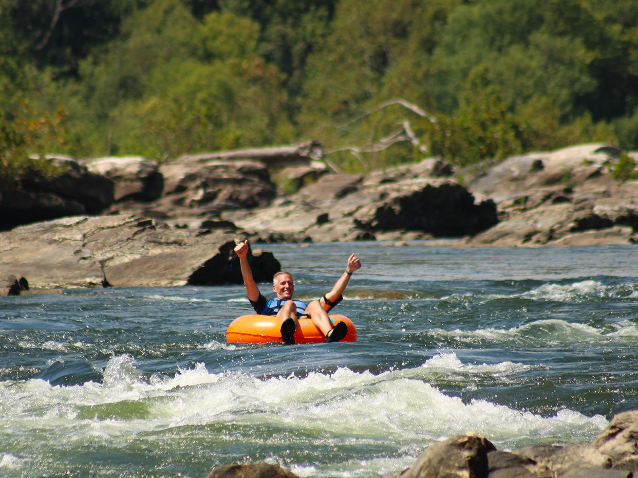 A person smiles and raises their arms while floating on an orange inner tube down a river with white water, surrounded by rocks and lush green trees.