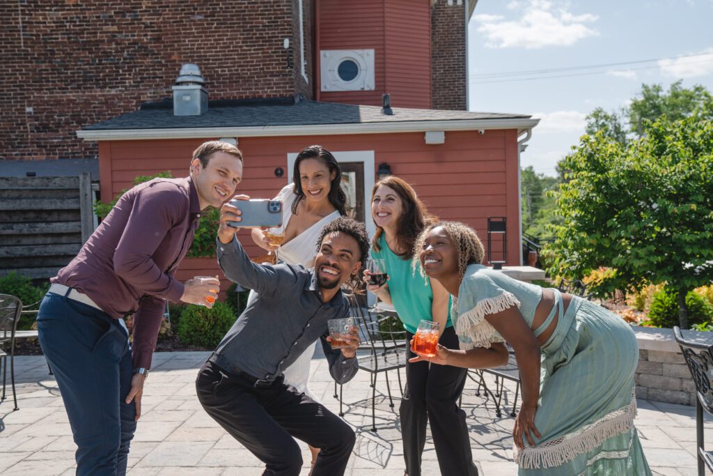 Five people stand together outdoors, smiling and posing for a selfie. Some of them hold drinks, and they are in front of a red building on a sunny day. There are trees and patio furniture in the background.