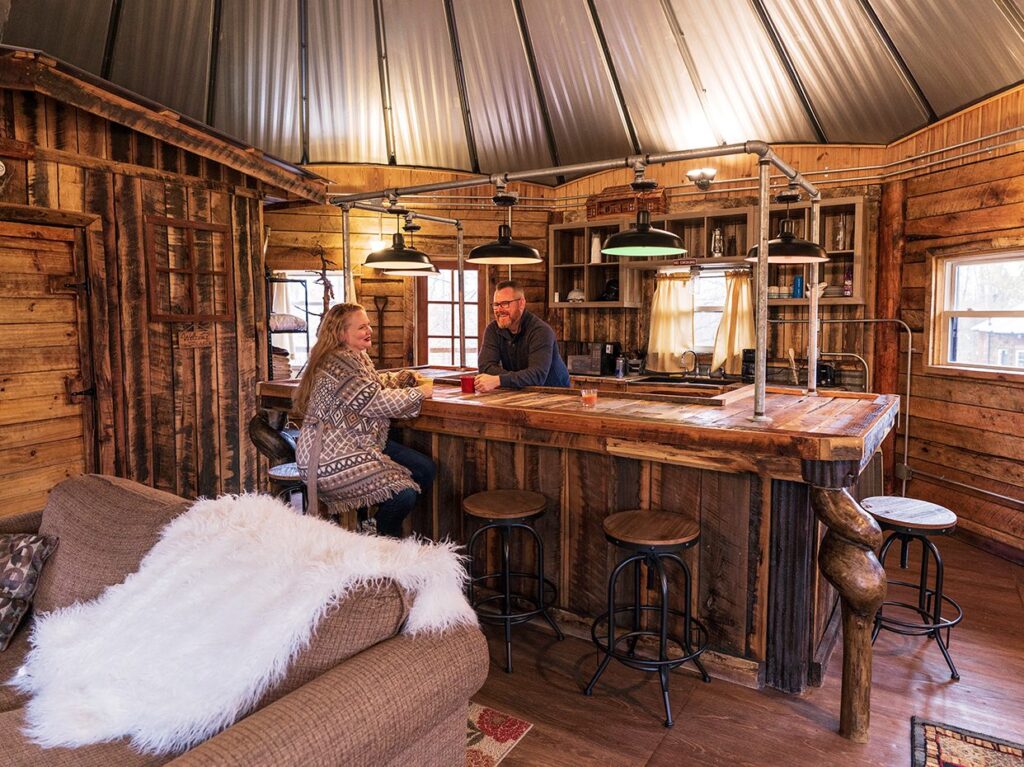 Two people sit at a rustic wooden bar inside a cozy cabin with wood-paneled walls, hanging lights, and a couch with a white blanket in the foreground. The atmosphere is warm and inviting.