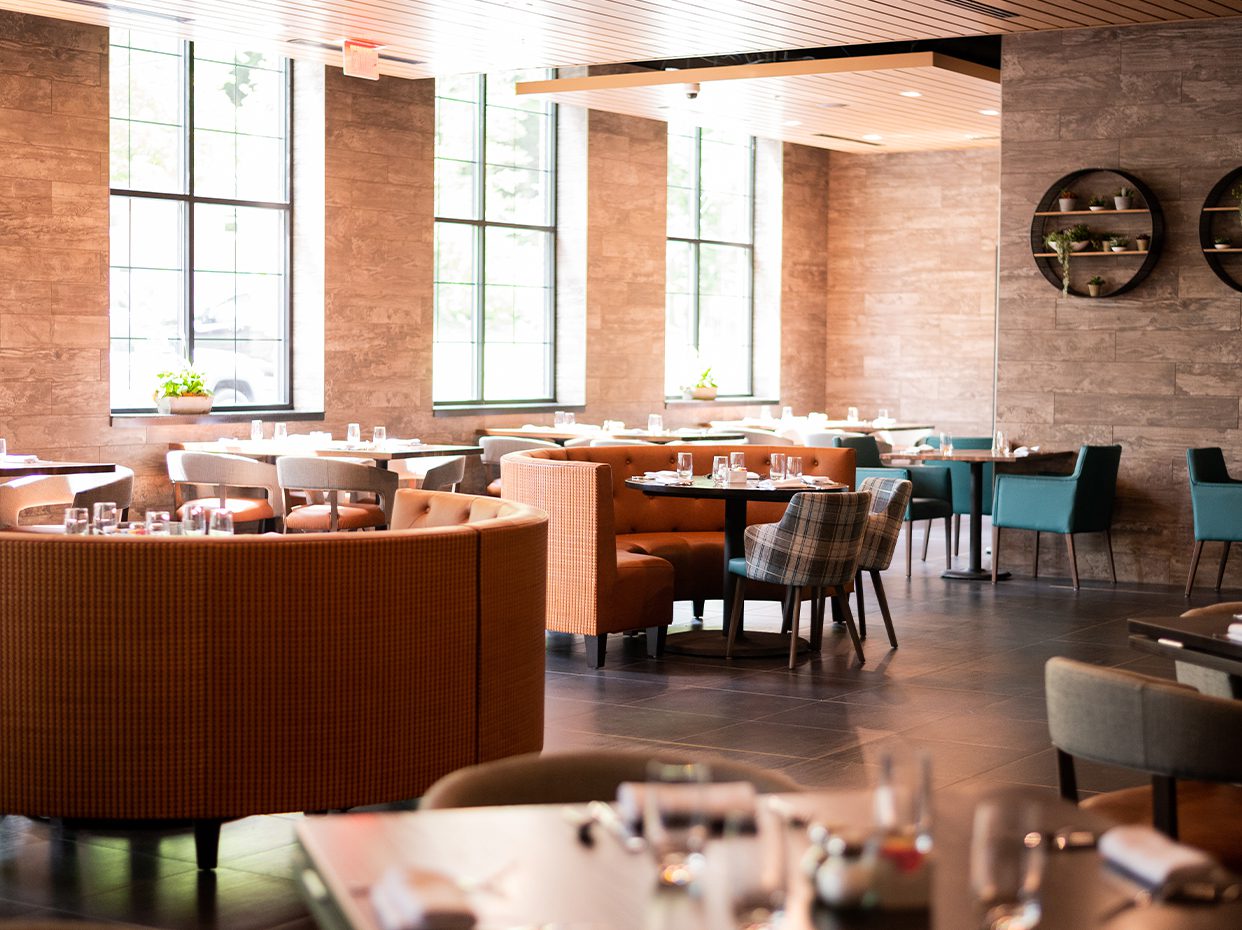 A modern, sunlit restaurant interior with large windows, orange and blue upholstered chairs, round and rectangular tables set with glassware, and neutral-toned walls decorated with round shelves holding plants.