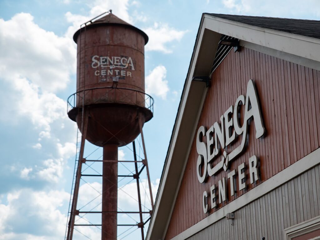 A rusty water tower and a building both display the words Seneca Center against a partly cloudy sky. The water tower is cylindrical with a conical top, and the building has red siding and a sloped roof.