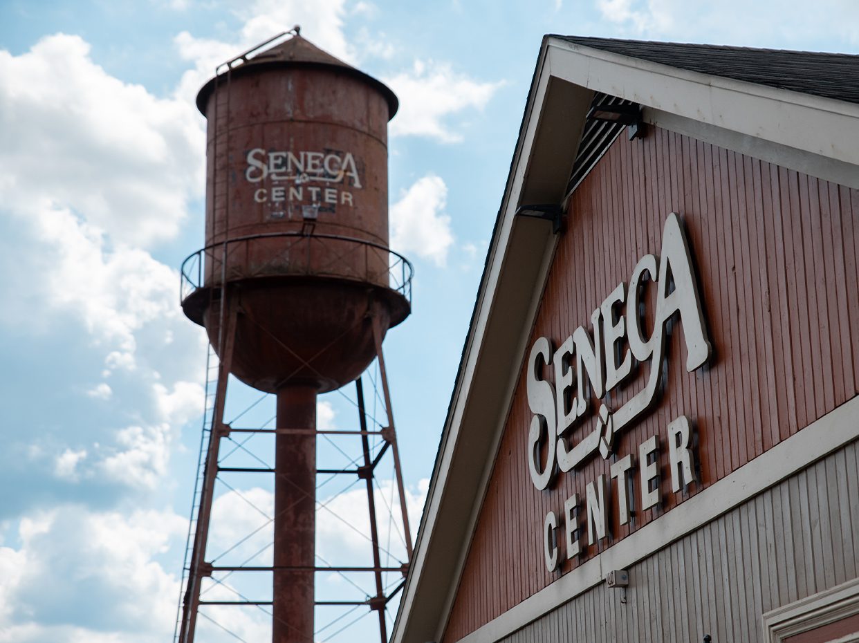 A rusty water tower and a building both display the words Seneca Center against a partly cloudy sky. The water tower is cylindrical with a conical top, and the building has red siding and a sloped roof.