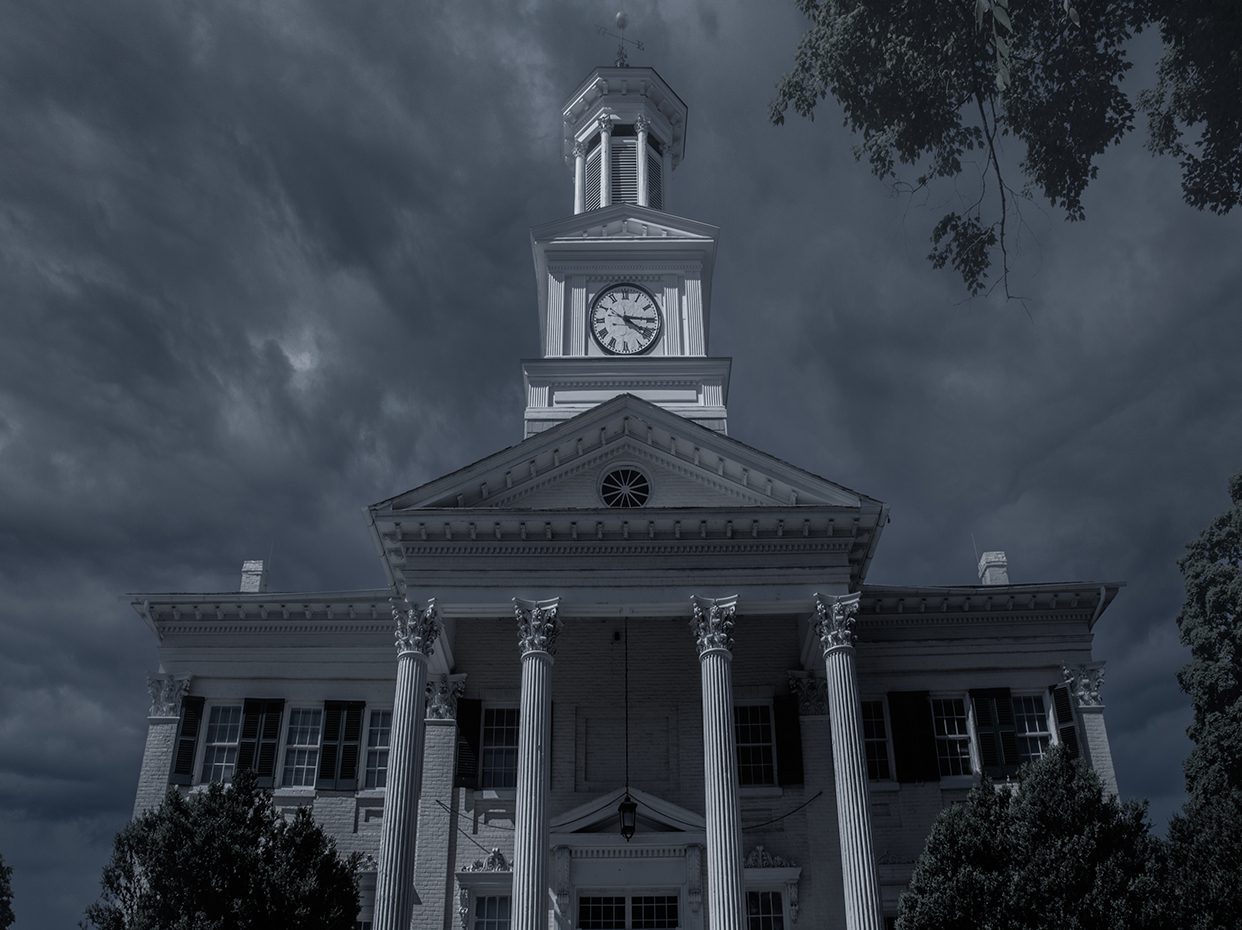 A large, white, columned building with a clock tower stands under a dark, cloudy sky, creating an eerie, dramatic atmosphere. Trees frame the scene on both sides.