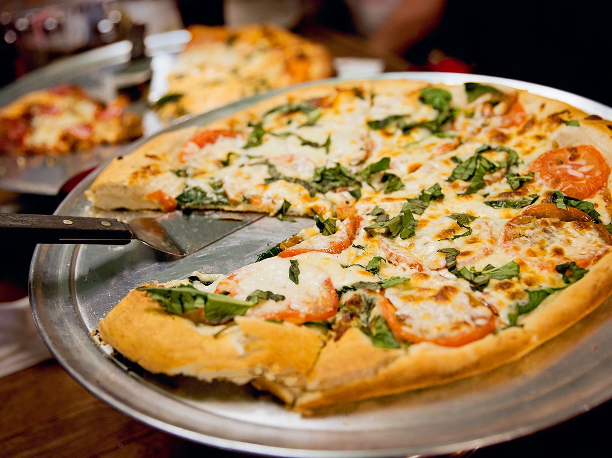 A close-up of a freshly baked pizza topped with tomatoes, spinach, and melted cheese on a metal tray. A pizza cutter is used to lift a slice. Another pizza is visible blurred in the background.