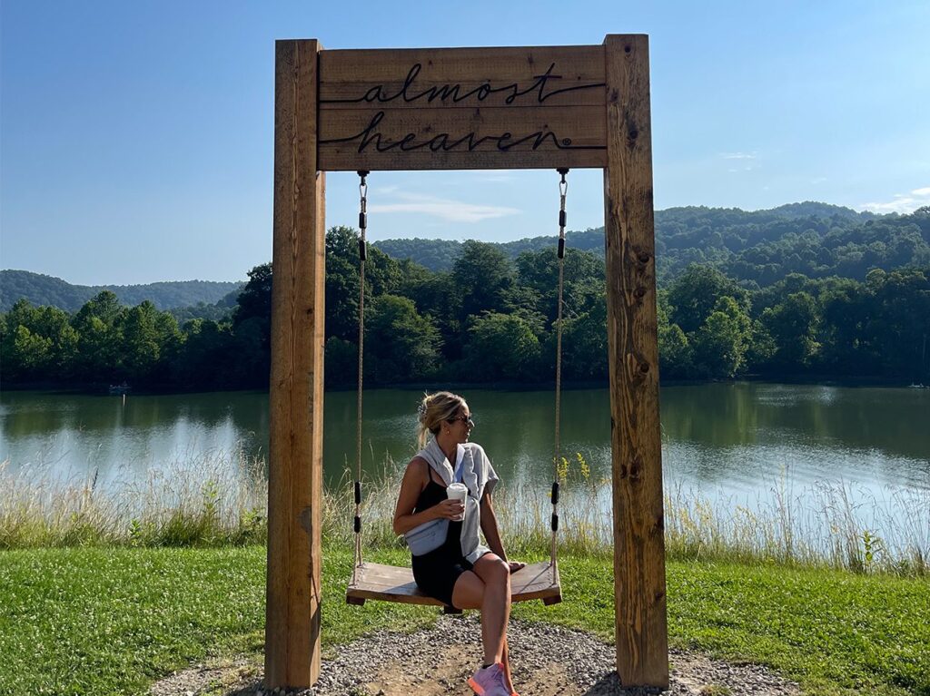 A woman sits on a swing under a wooden sign reading almost heaven, by a lake with forested hills in the background on a sunny day.