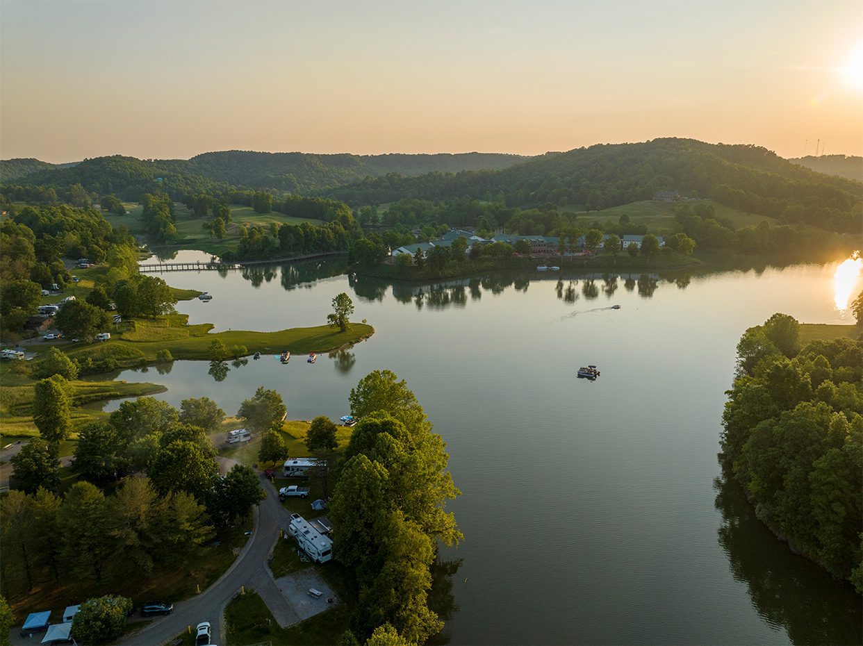 A tranquil lake surrounded by lush green trees and hills at sunset, with a few boats on the water, a bridge in the distance, and RVs parked along the shoreline.