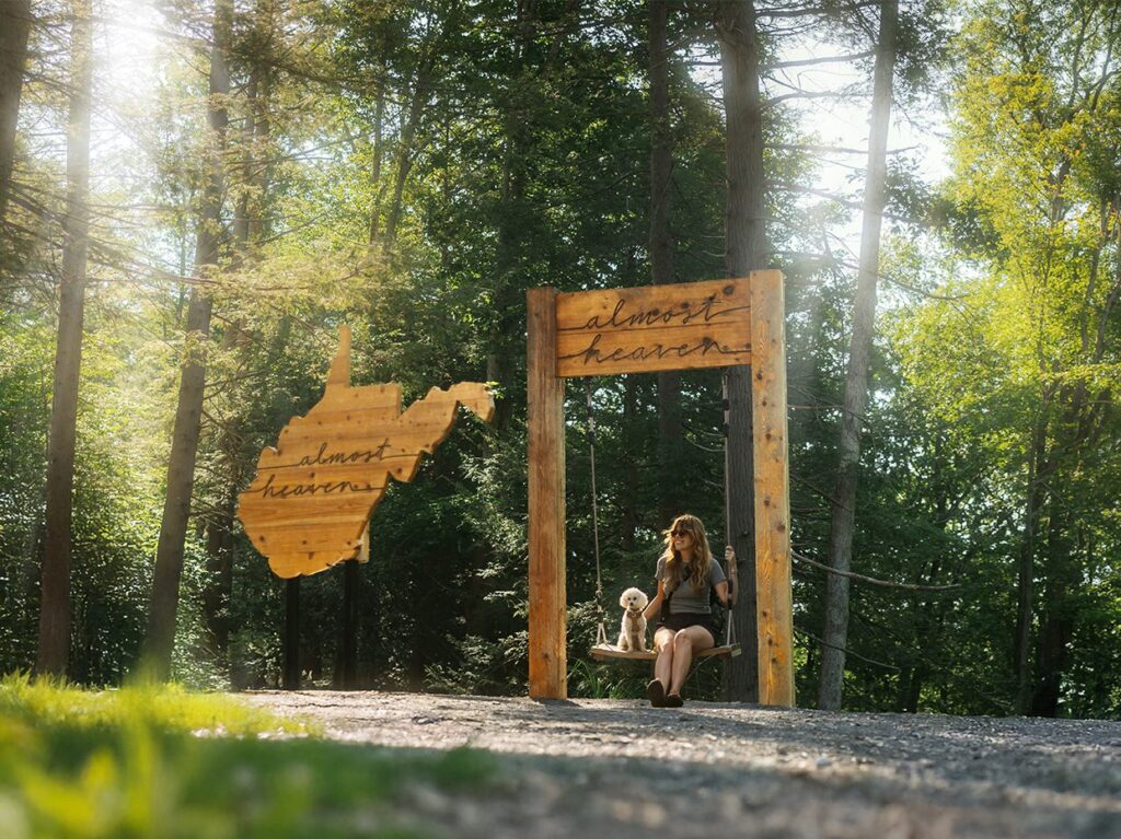 A woman sits on a swing holding a small dog in a sunny, wooded area. Next to her is a wooden sign shaped like West Virginia with almost heaven written on both the sign and swing frame.