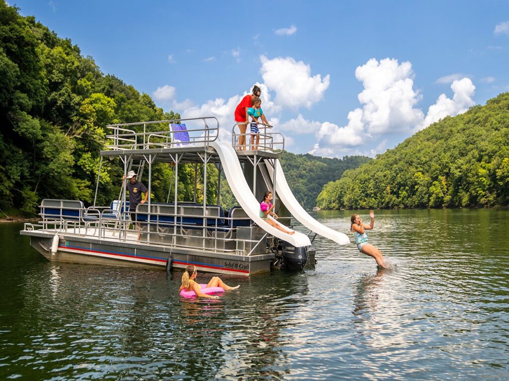 A pontoon boat with slides is anchored on a calm lake surrounded by forested hills. Adults and children enjoy the water, swimming, and sliding into the lake under a bright blue sky with scattered clouds.