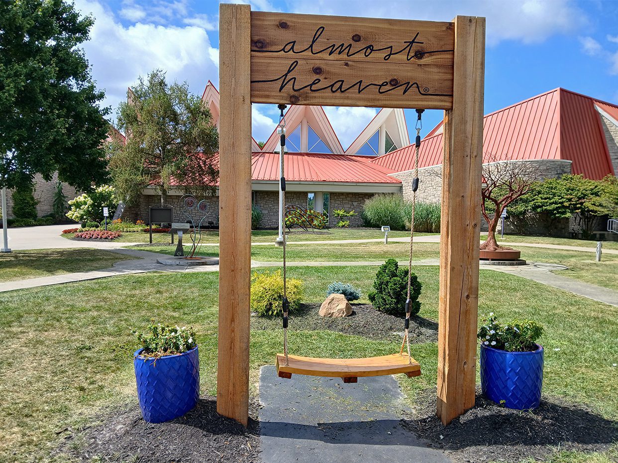 A wooden swing set with almost heaven written on top stands between two blue planters with flowers, in front of a modern building with red rooftops and a landscaped garden.