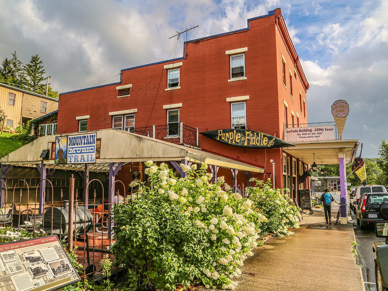 A red brick building houses the Purple Fiddle café and Mountain Trail store, with a patio, blooming white flowers, and a man walking on the wet sidewalk under a cloudy sky.
