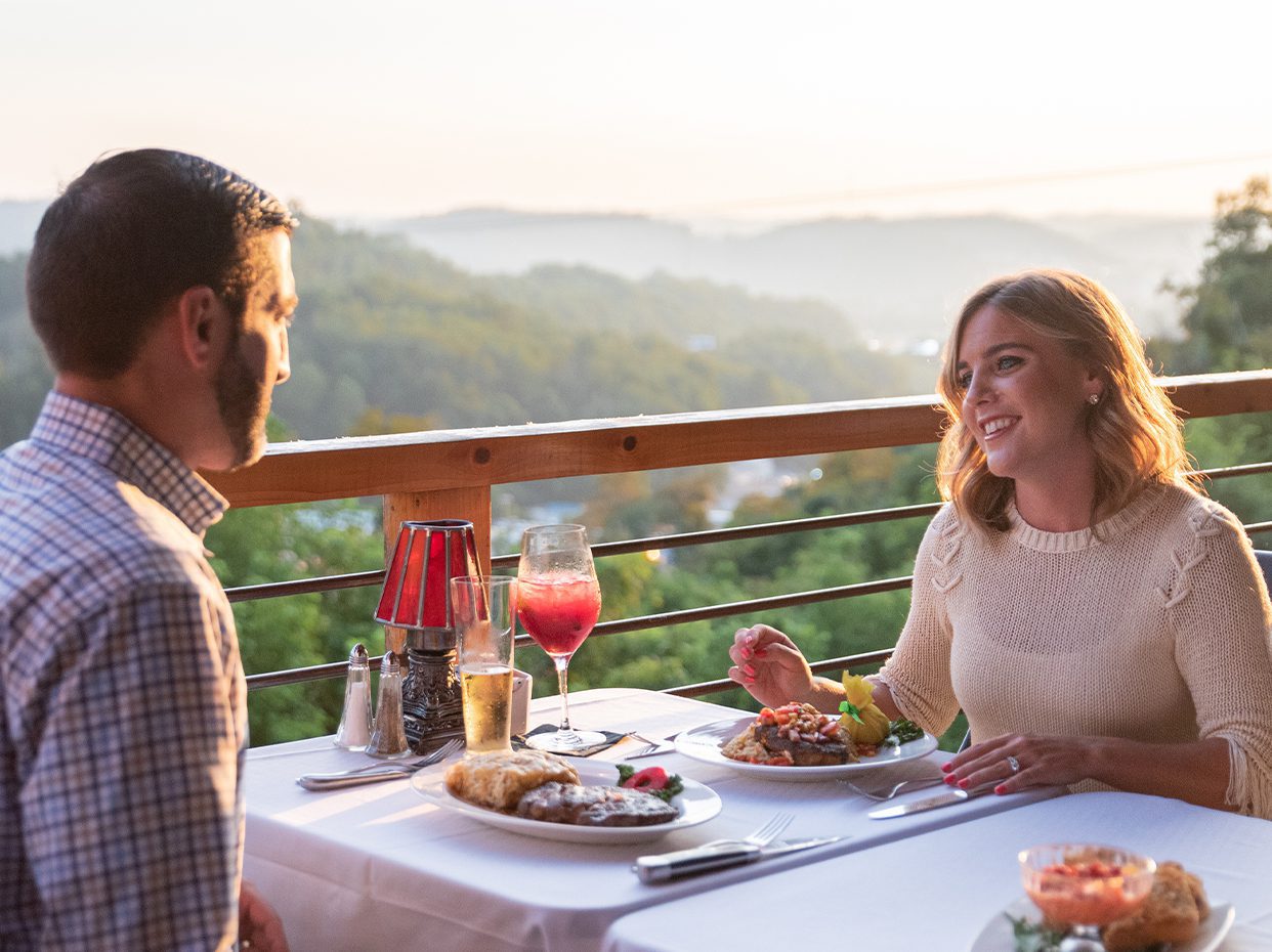 A man and a woman dine outdoors at a table set with food and drinks, overlooking a scenic, sunlit landscape with rolling hills in the background. The woman is smiling and talking to the man.