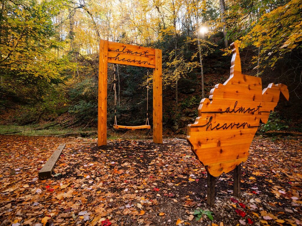 A wooden swing labeled almost heaven stands in a forest with autumn leaves. Nearby, a wooden cutout of West Virginia also reads almost heaven among colorful trees and dappled sunlight.
