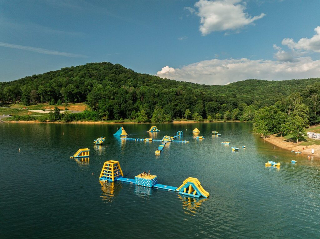 A floating inflatable water park with slides and obstacles sits on a lake, surrounded by forested hills under a partly cloudy sky. People are playing on the inflatables and along the sandy shoreline.