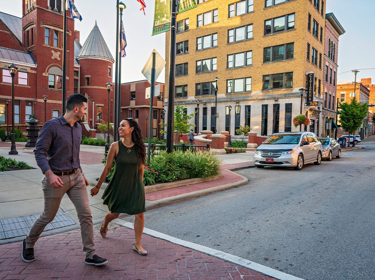 A man and woman walk hand-in-hand across a crosswalk in a city downtown area, surrounded by historic brick buildings, greenery, and parked cars on a sunny day.