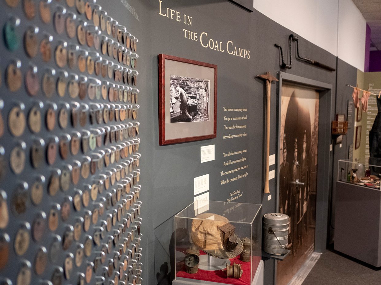 Museum exhibit wall titled “Life in the Coal Camps,” displaying a grid of numbered metal tags, mining tools, vintage photos, and artifacts related to coal mining life.