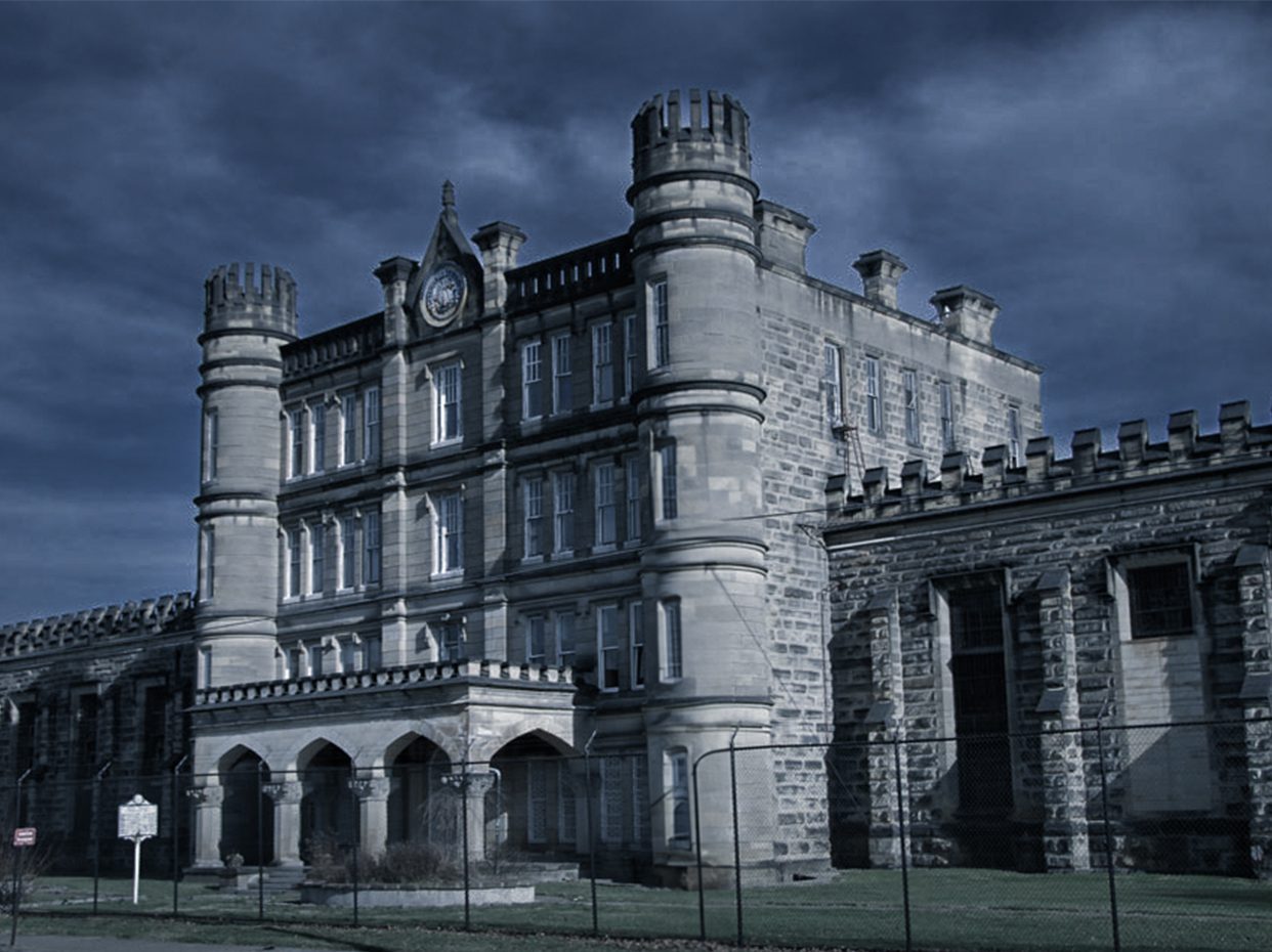 A large, old stone building with castle-like towers and arched entrances, surrounded by a fence, sits under a cloudy, moody sky.