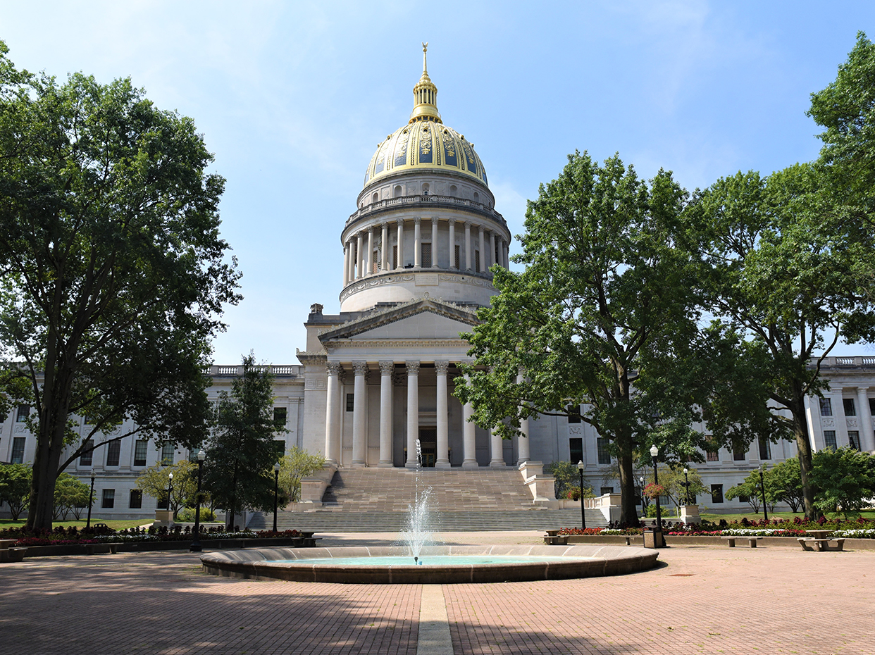 A large domed government building with tall columns and a gold-topped dome, framed by trees, stands behind a circular fountain and brick courtyard under a clear sky.