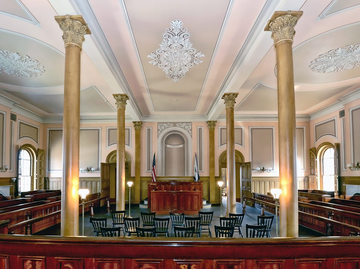Elegant, historic courtroom with ornate ceiling, tall golden columns, wood-paneled walls, American flags, judge’s bench, empty wooden chairs in the center, and large arched windows letting in natural light.