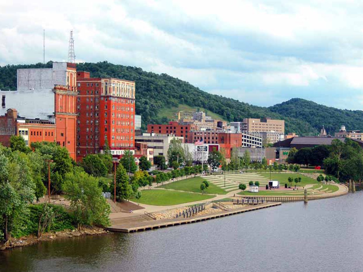 A riverfront park with green lawns and walkways lines the water’s edge, with red brick buildings and other city structures in the background, set against tree-covered hills under a cloudy sky.