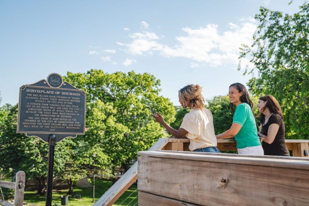 Three women stand on a wooden platform outdoors, reading a historic marker titled Birthplace of Bourbon. Lush green trees and a bright blue sky are in the background.