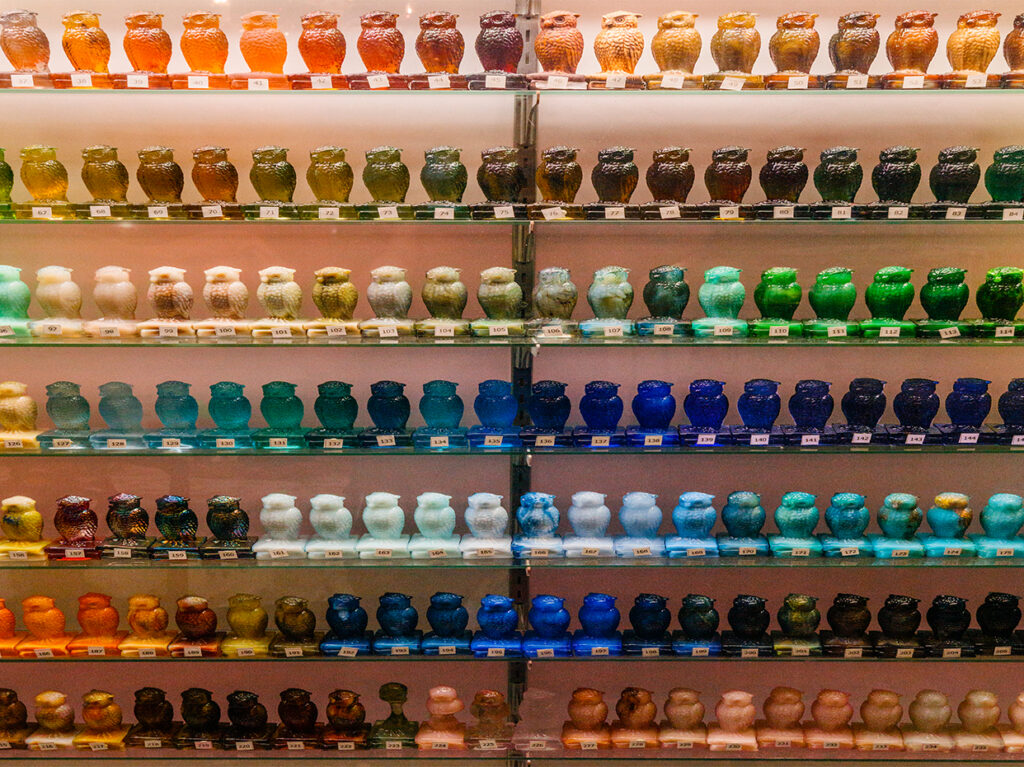 Shelves filled with ceramic owl figurines arranged in rows by color gradient, from warm reds and yellows at the top to cooler blues and greens, and ending with browns and neutral tones at the bottom.