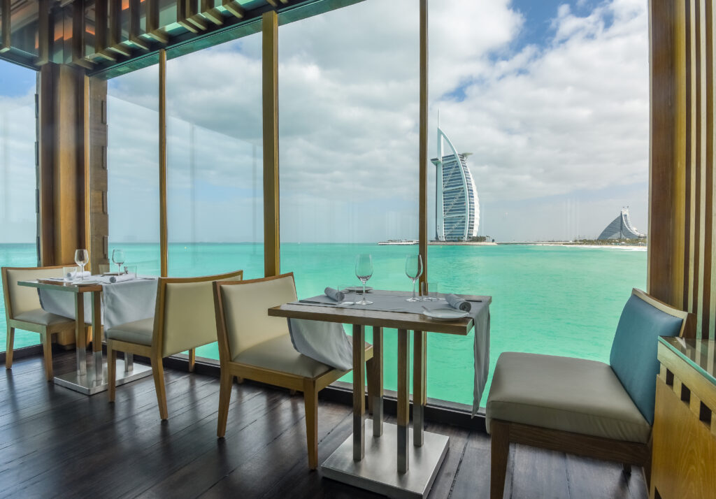 Modern restaurant with tables set by large windows overlooking turquoise water and a distant view of the Burj Al Arab and another building under a partly cloudy sky.