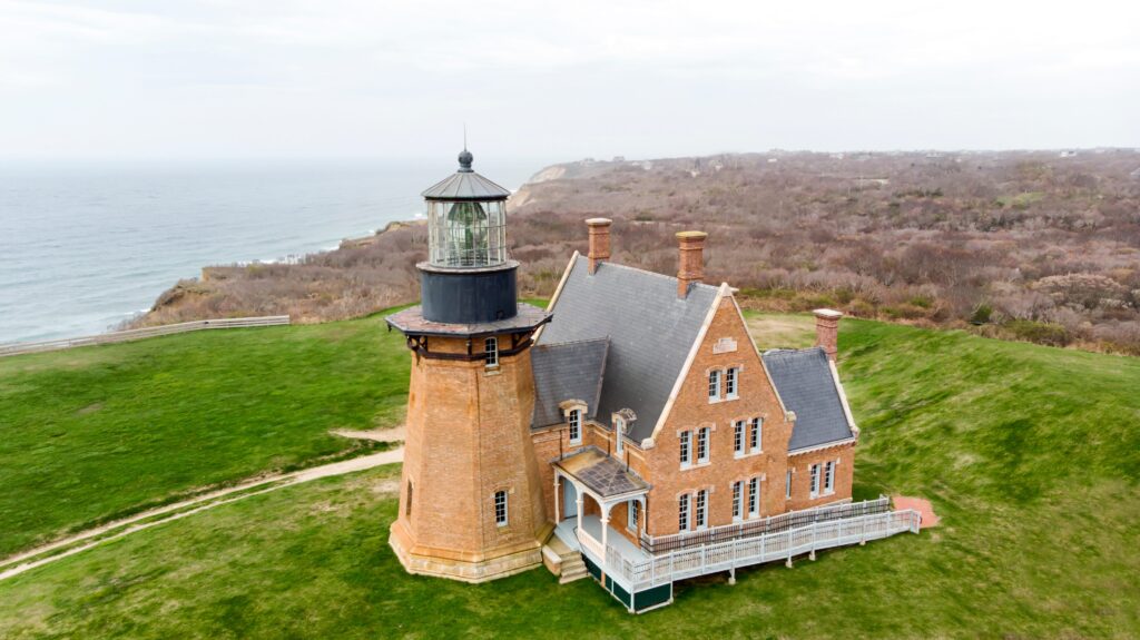 Aerial view of a brick lighthouse with an attached house, surrounded by green grass on a cliff overlooking the ocean. The sky is overcast and the distant shoreline is visible.