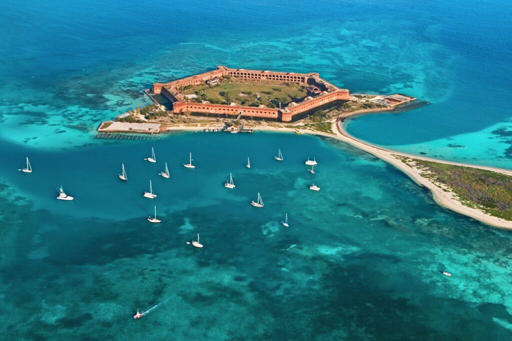 Aerial view of a historic brick fort surrounded by turquoise water, connected to a sandy strip with lush greenery, and several white sailboats anchored nearby in the clear blue sea.