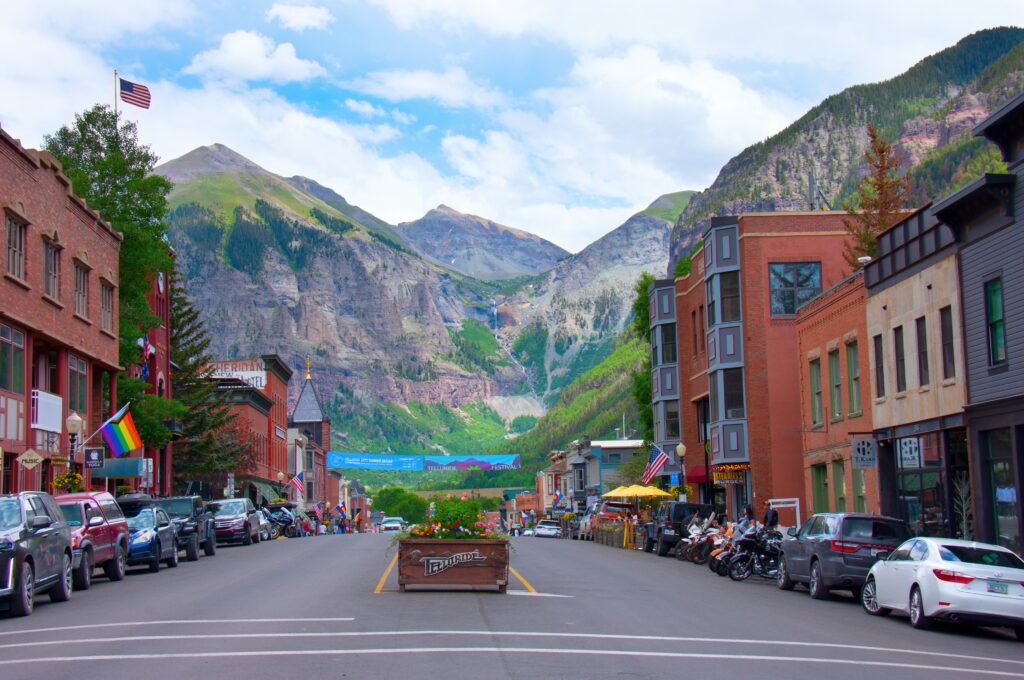 A colorful small town street lined with cars, shops, and flags, set against a backdrop of tall, green mountains under a partly cloudy sky.