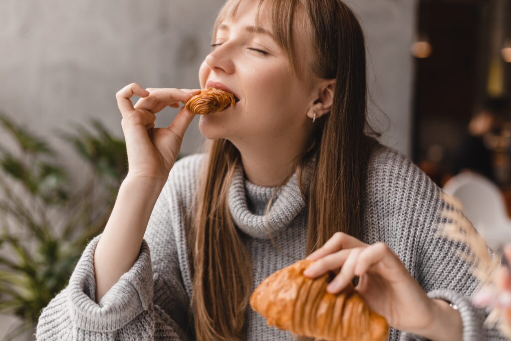 A woman with long hair and a gray sweater sits indoors, eyes closed, enjoying the taste of a croissant, holding another piece in her hand.