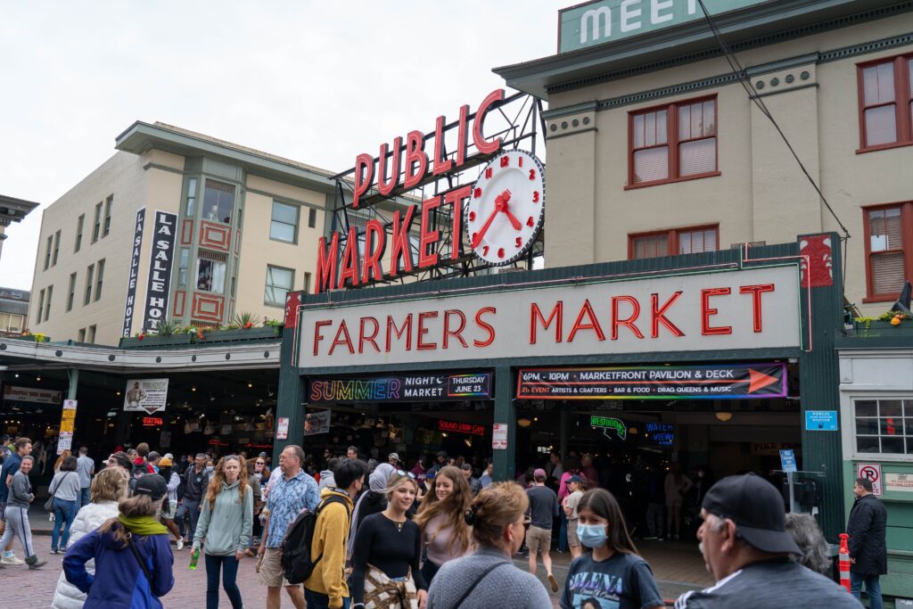 Crowds walk past the iconic neon sign for Pike Place Market in Seattle, with “Public Market” and “Farmers Market” prominently displayed. People in casual clothing fill the bustling outdoor market scene.