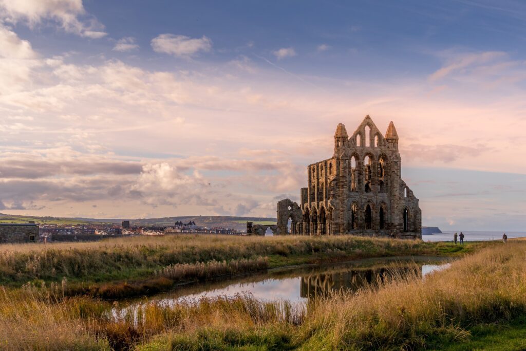 Ruins of Whitby Abbey stand on a grassy hill at sunset, reflected in a nearby pond, with a few people walking in the distance and a town and cloudy sky in the background.