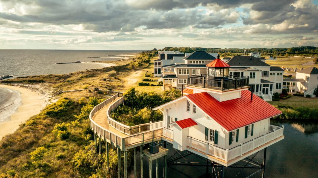 Aerial view of a white lighthouse with a red roof on stilts, connected to the beach by a wooden boardwalk, surrounded by modern houses and overlooking the coastline under a partly cloudy sky.