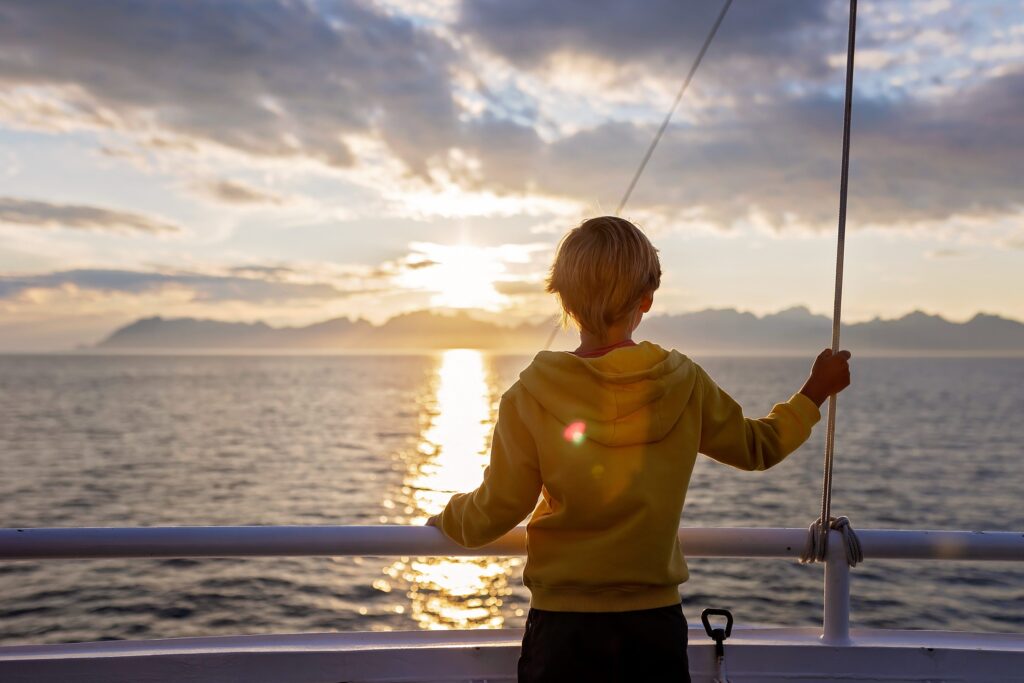 A child in a yellow hoodie stands on a boat, holding a railing and looking out at the ocean during sunset, with mountains and clouds in the distance and sunlight reflecting on the water.
